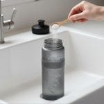 Mano añadiendo percarbonato con una cuchara a una botella reutilizable para aplicar un método de limpieza natural y eliminar el mal olor.